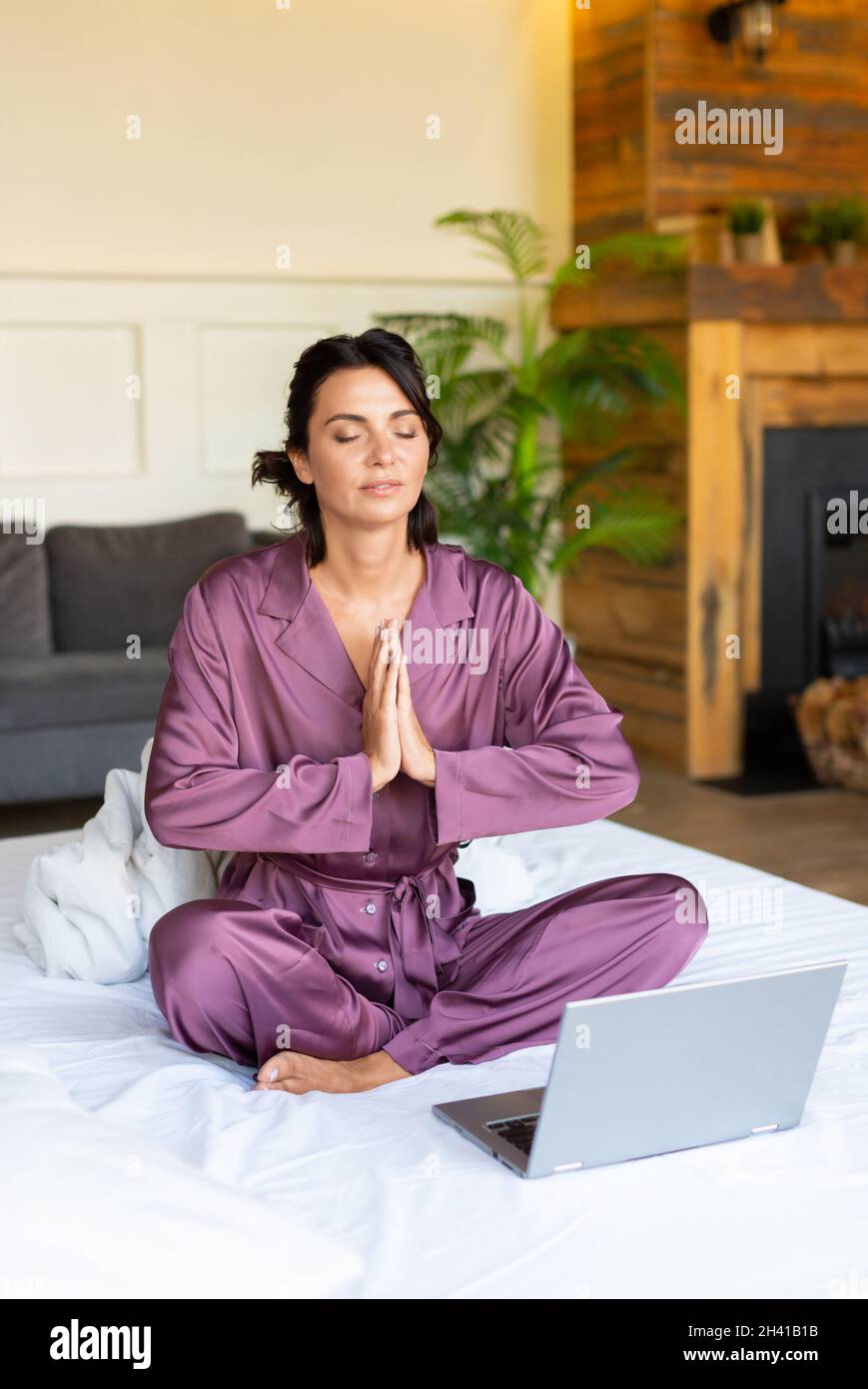 Concept of restoring strength and mental energy through yoga practice - middle aged woman meditating in bed in front of laptop monitor. Vertical photo Stock Photo