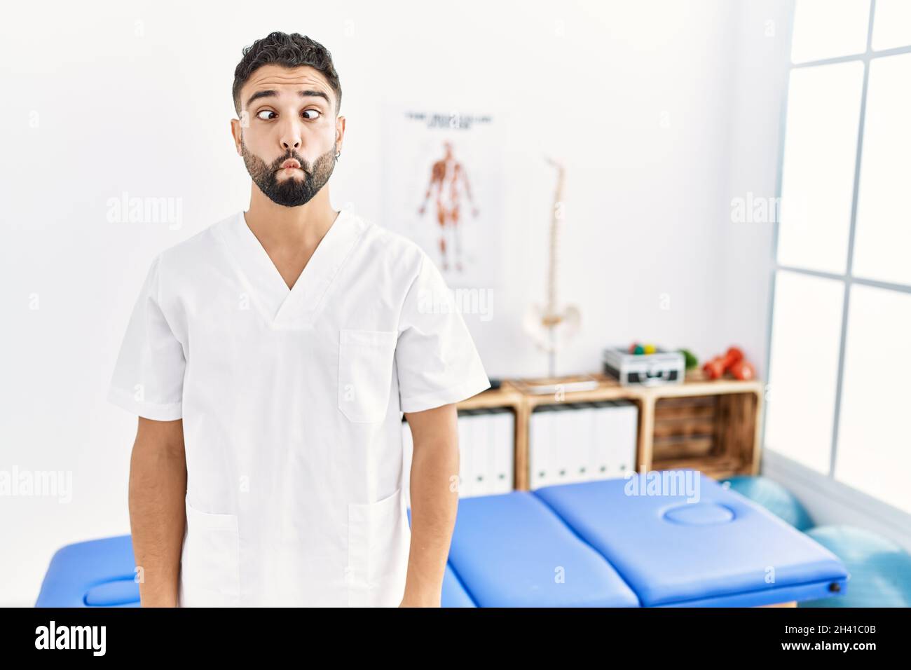 Young handsome man with beard working at pain recovery clinic making fish face with lips, crazy and comical gesture. funny expression. Stock Photo