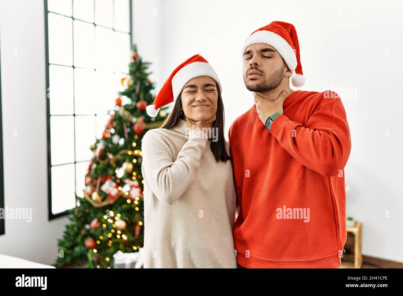 Young hispanic couple standing by christmas tree touching painful neck, sore throat for flu