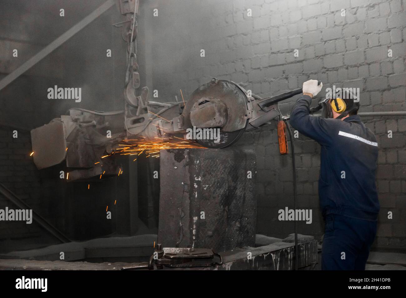 A man in a working jumpsuit and respirator, cleans cast iron tubing with heavy equipment with a grinding stone in the workshop of an industrial plant. Stock Photo