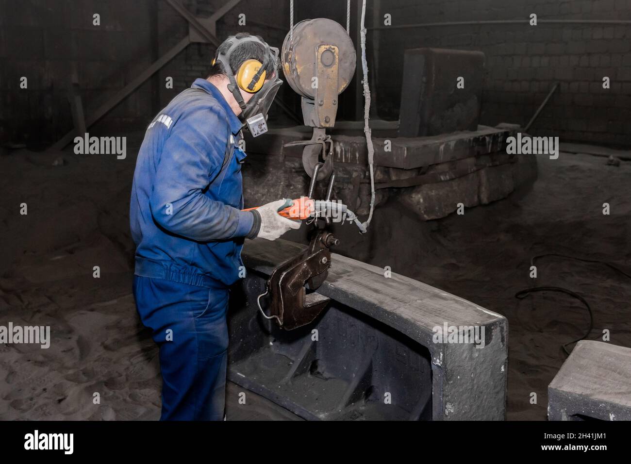 A working man in overalls and a respirator holds in his hand the control panel of the lifting mechanism in front of the cast iron concrete tube, in th Stock Photo
