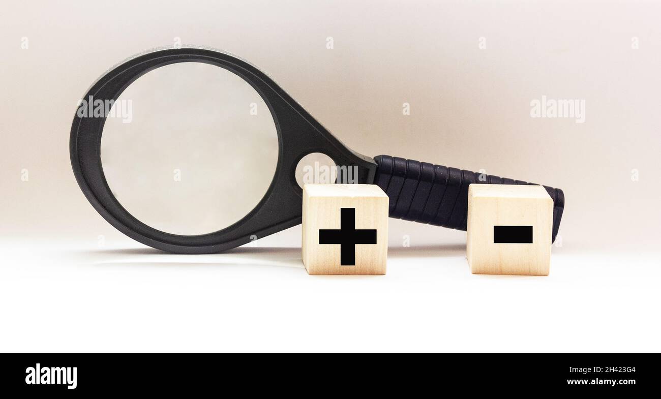 Two wooden cubes with mathematical symbols plus and minus, near a magnifying glass on a white background Stock Photo