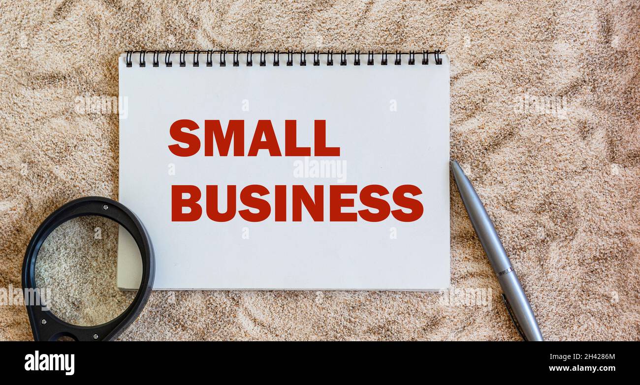 The text SMALL BUSINESS is written on a notebook and a wooden table, next to it lies a magnifying glass and a pen Stock Photo
