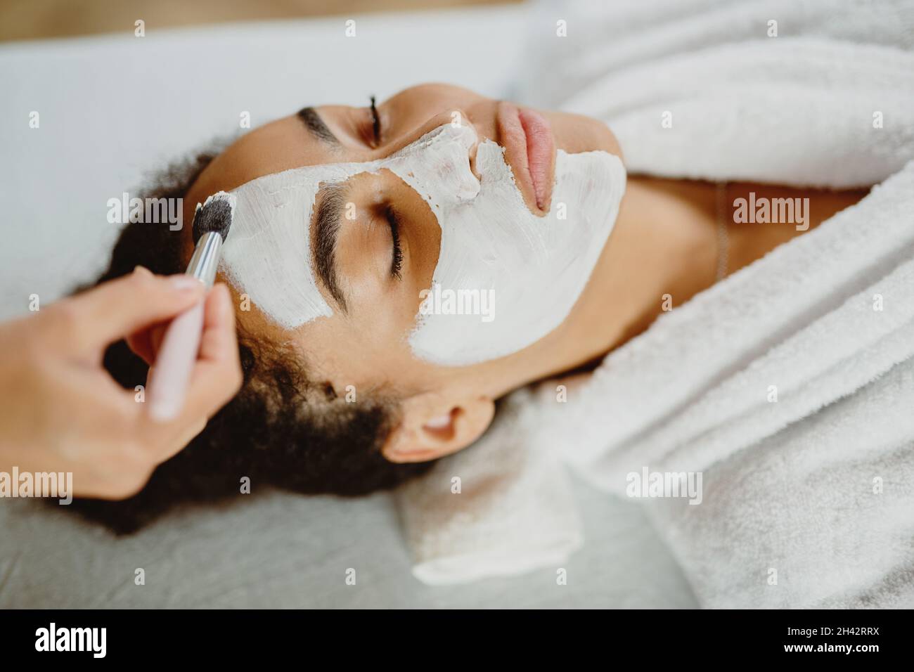 Beautician applies white clay face mask with a brush on a woman's face. Stock Photo