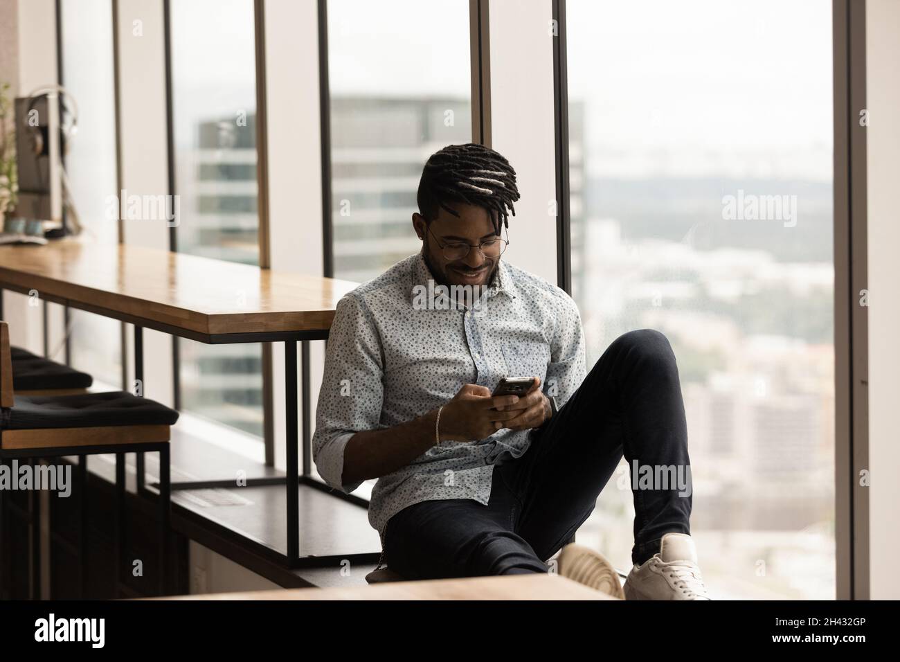 Relaxed african ethnicity man using smartphone, sitting on windowsill. Stock Photo