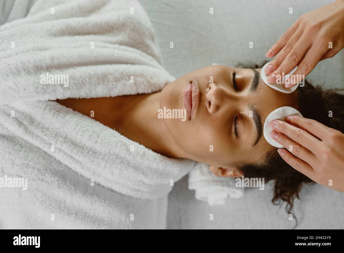 Professional beautician cleansing skin with cotton pads to dark-skinned young woman. Stock Photo
