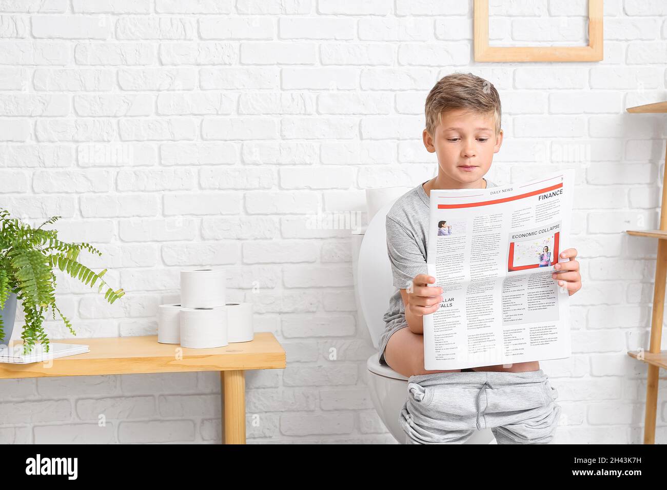 Little boy reading newspaper while sitting on toilet bowl in bathroom Stock Photo