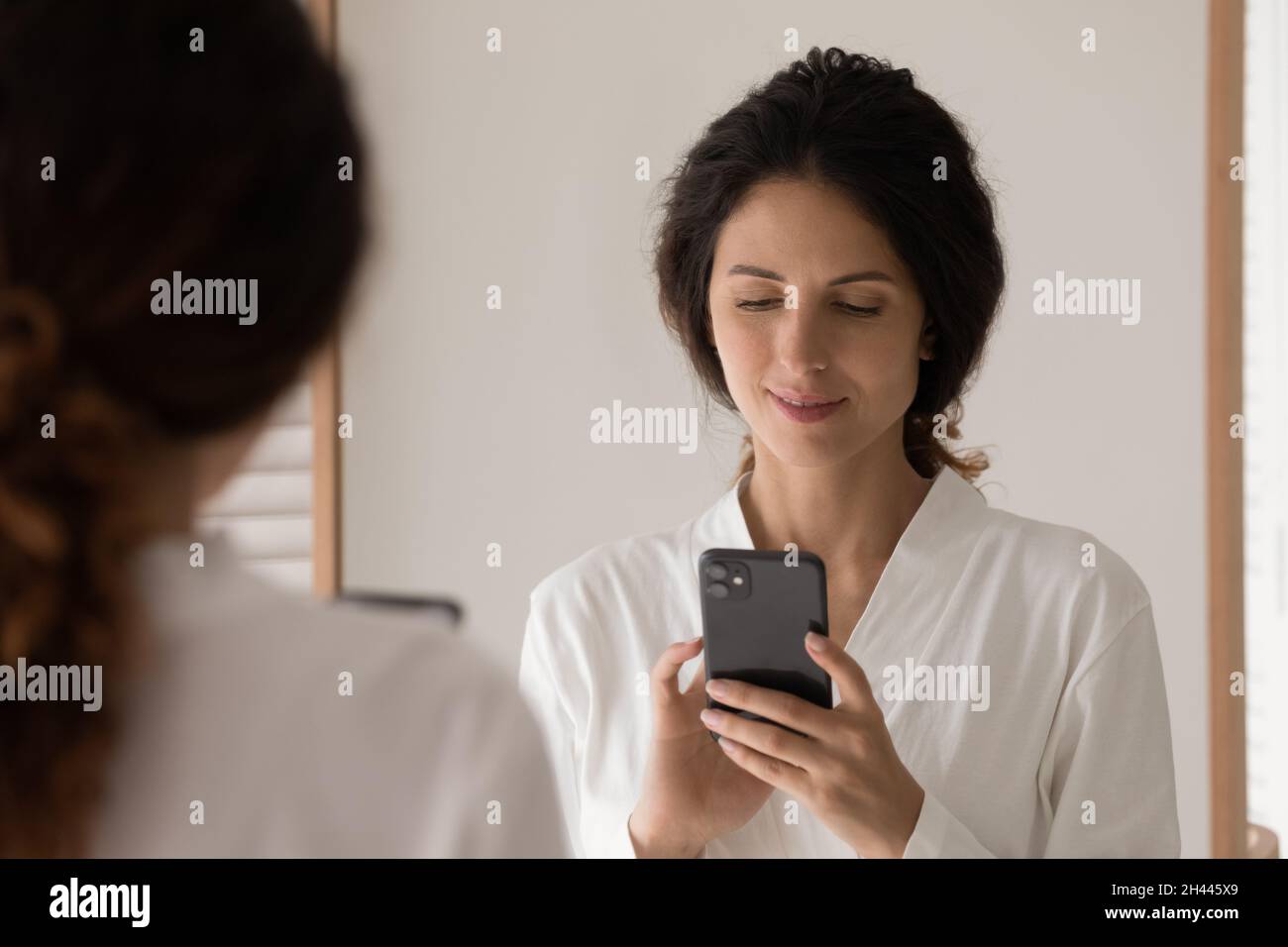 Hispanic woman wear bathrobe stands in bathroom with smartphone Stock Photo