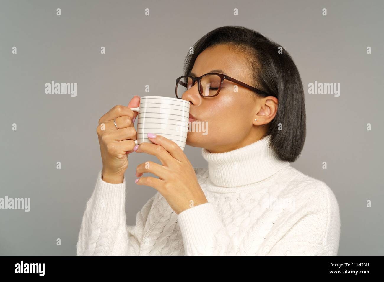 African woman in cozy sweater drinking hot coffee or tea with closed eyes. Winter drinks concept Stock Photo