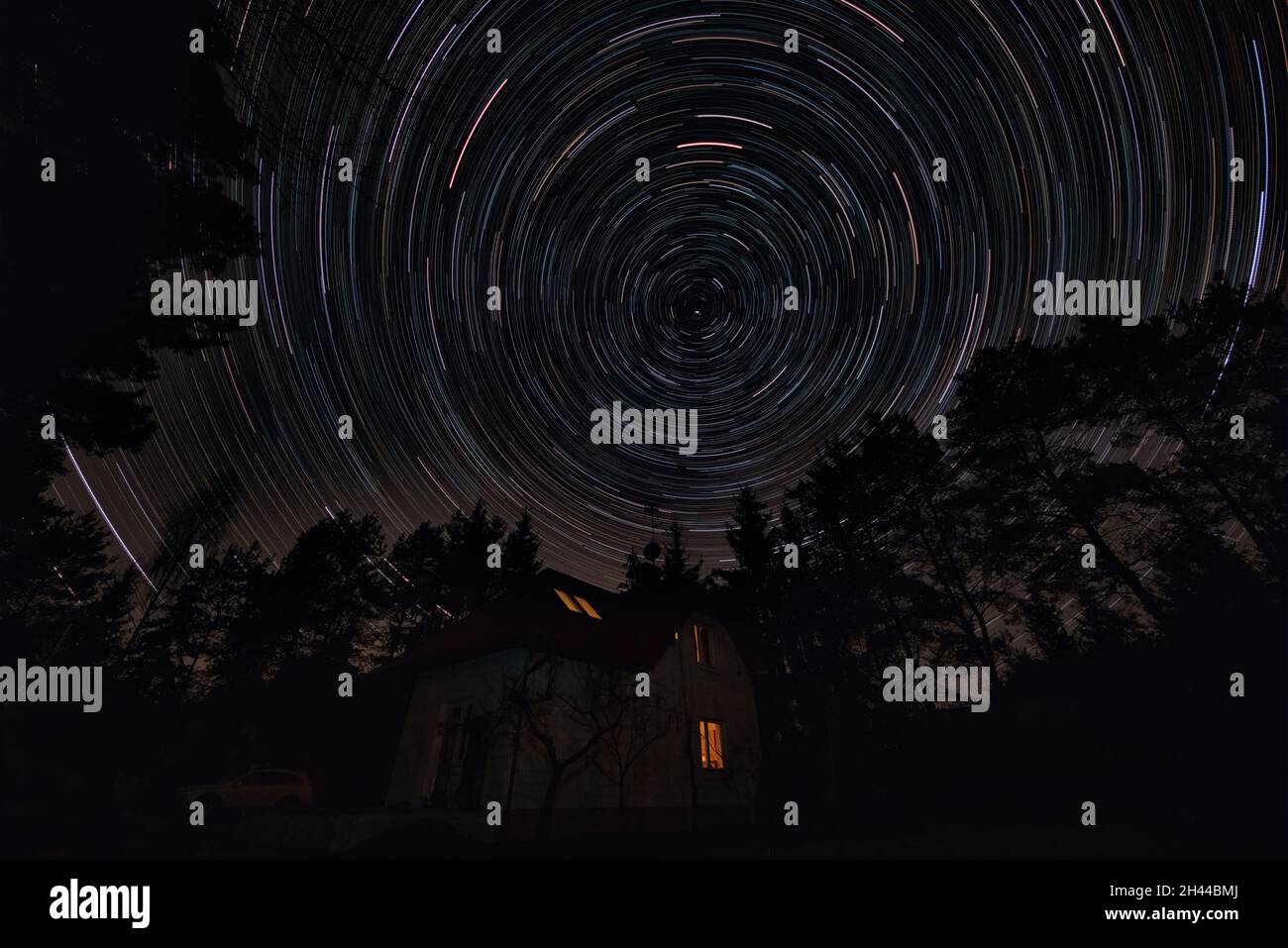 House surrounded by trees under dark night sky. Movement of stars around pole star on north hemisphere. Startrails on night sky, long exposure composi Stock Photo