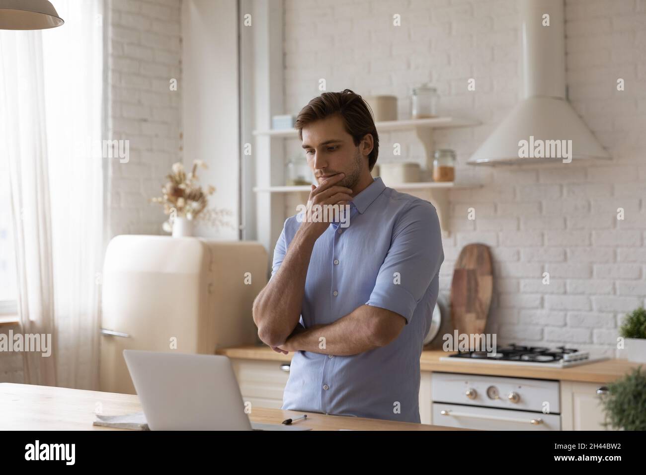 Man standing in kitchen staring at laptop thinks over task Stock Photo