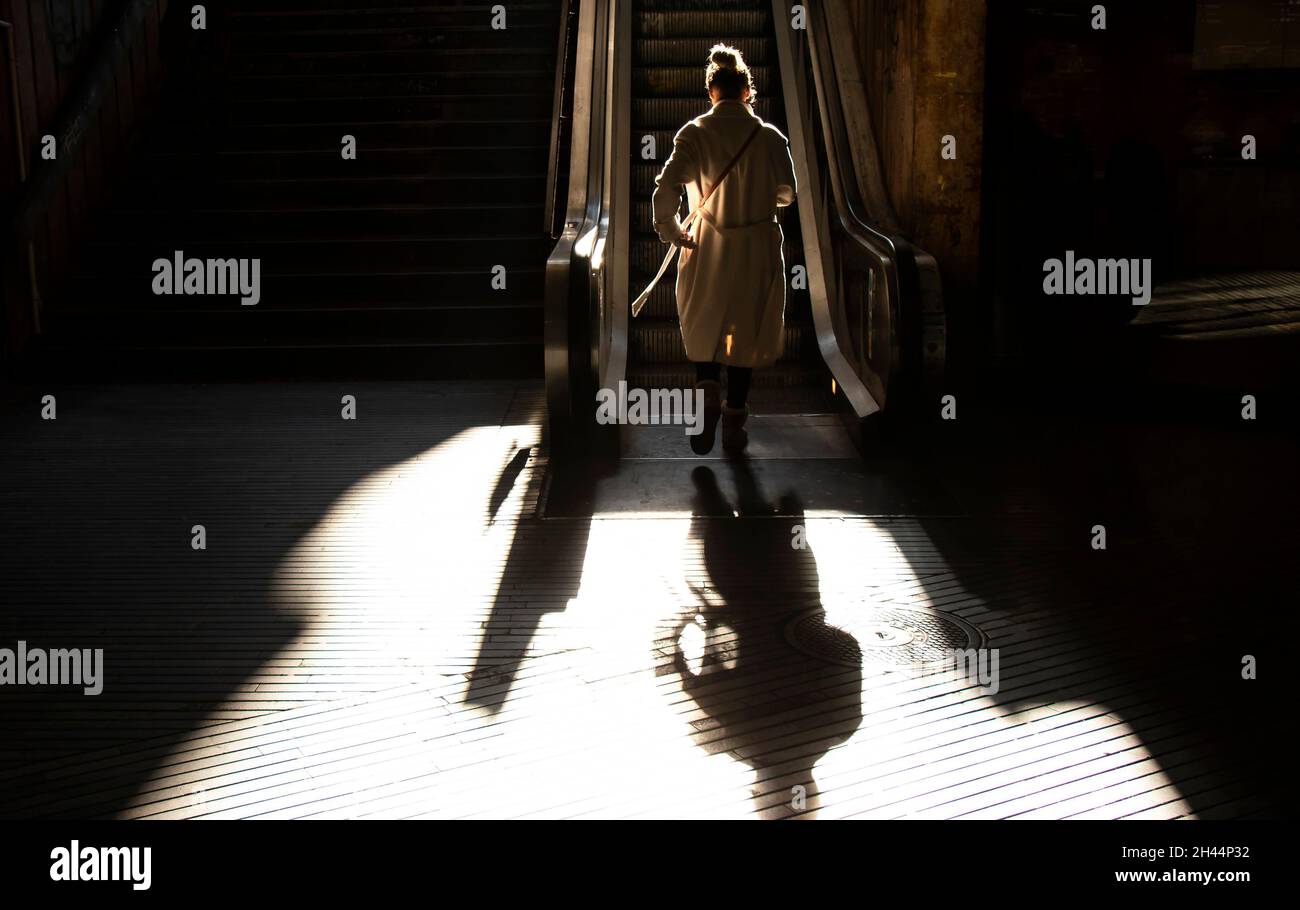 Belgrade, Serbia - October 16, 2020: One young woman in coat walking alone up the escalator public stairs in backlight sunlight, from behind, with sha Stock Photo