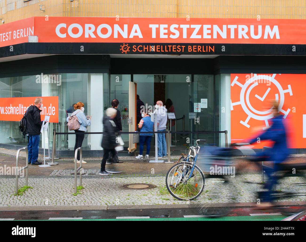 Berlin, Germany. 30th Aug, 2021. 30.08.2021, Berlin. People queue up and wait with masks in front of their faces in front of a Corona test centre in Steglitz to get tested. In the foreground a cyclist rides by. (Wiping effect due to longer exposure time). Credit: Wolfram Steinberg/dpa **Use in full format only**. Credit: Wolfram Steinberg/dpa/Alamy Live News Stock Photo