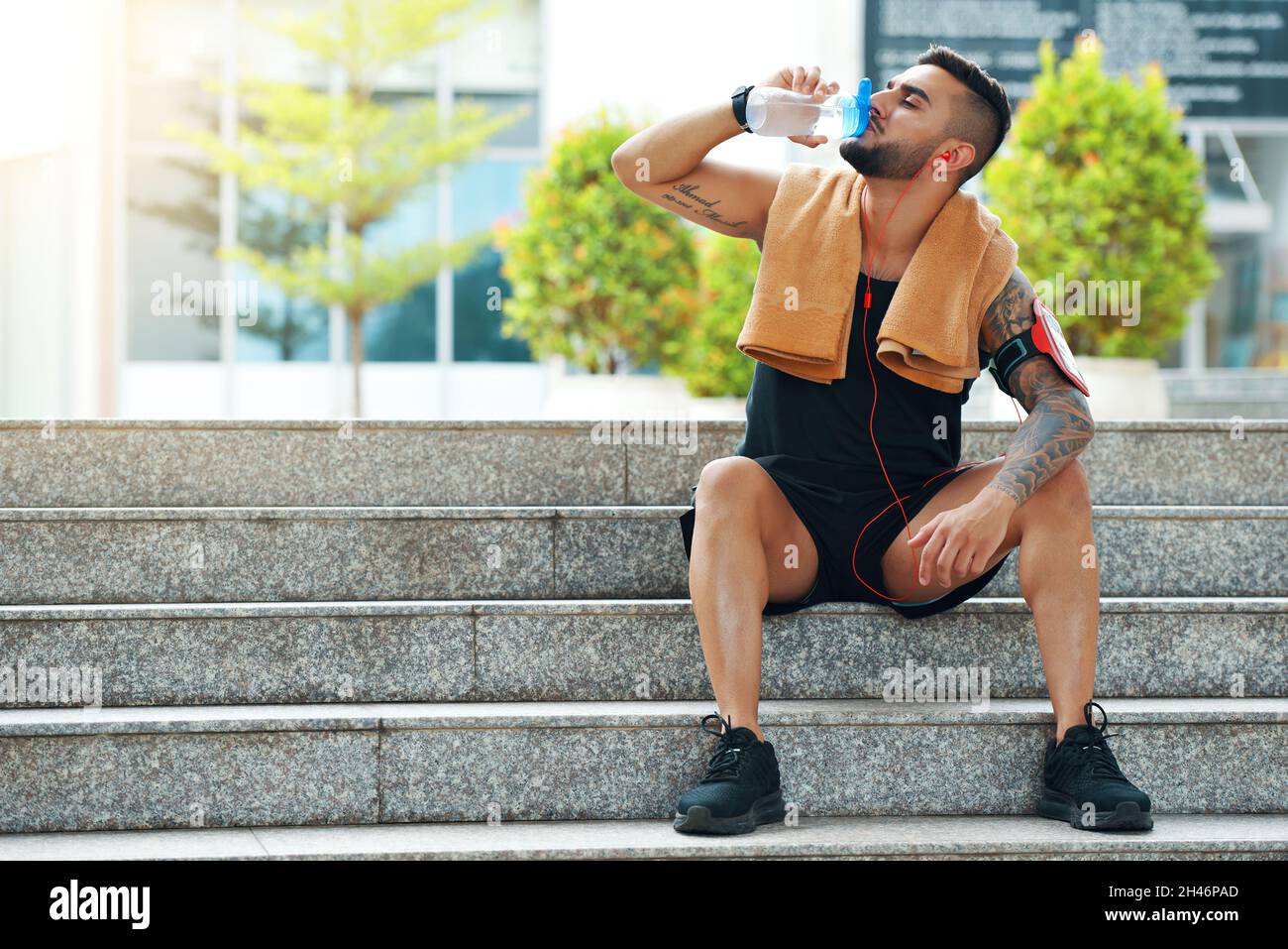 Fit handsome young man sitting on steps and drinking water after training outdoors Stock Photo