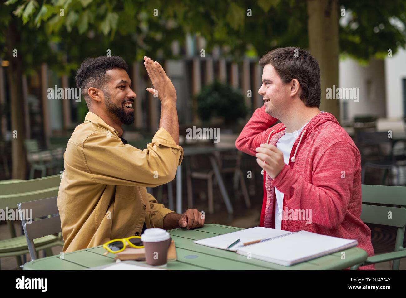 Young man with Down syndrome with mentoring friend sitting outdoors in cafe celebrating success. Stock Photo