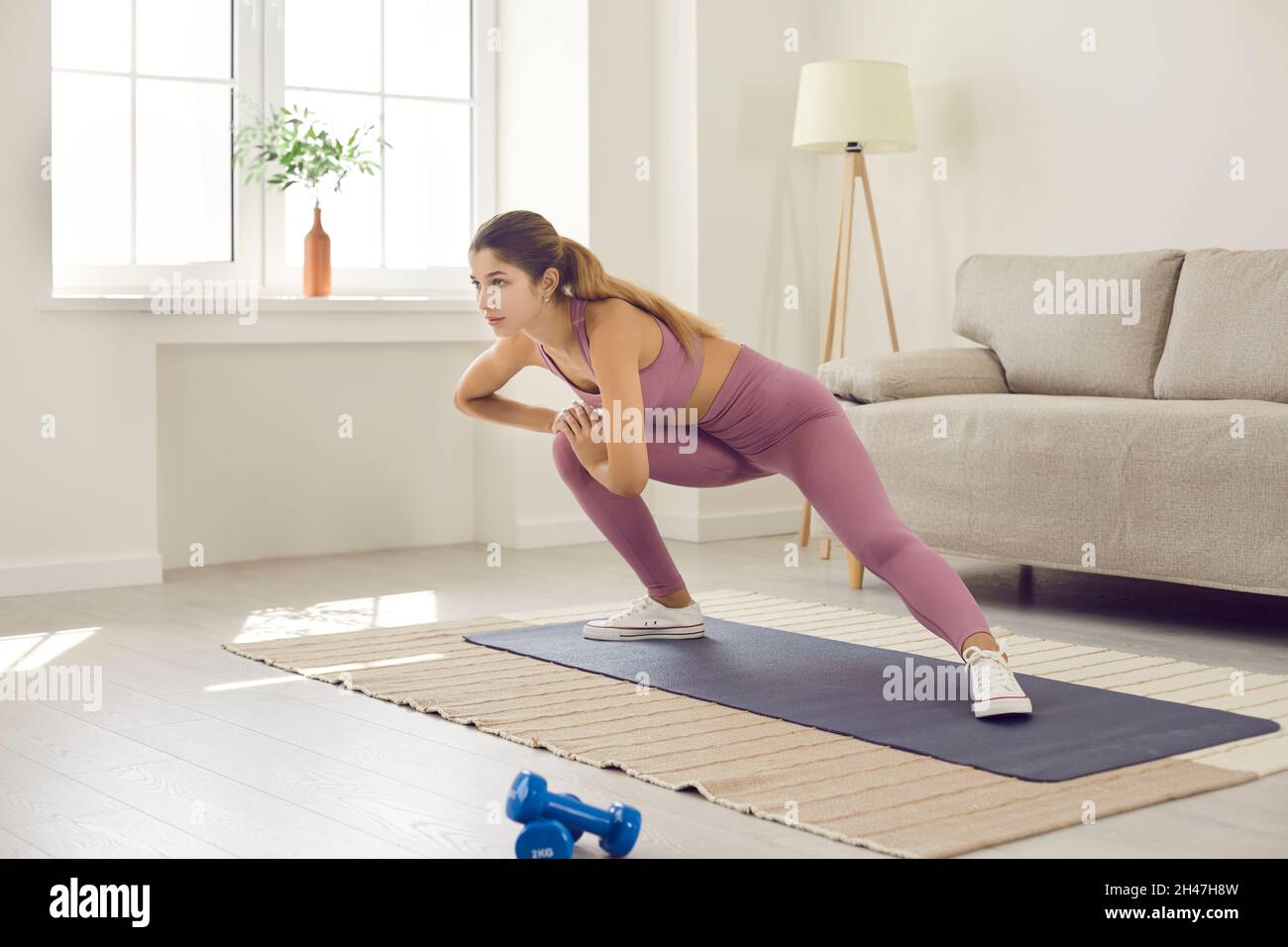 Young slender woman trains at home on sports mat and does stretching in room with morning sunlight. Stock Photo