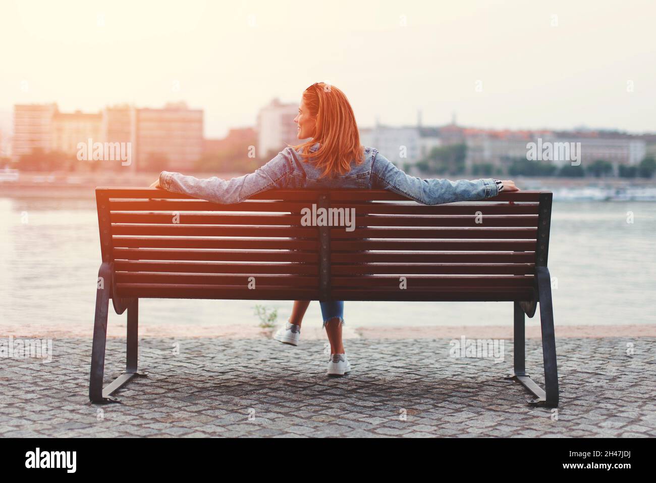Young redhead woman relaxing on bench at riverside in orange sunrise Stock Photo