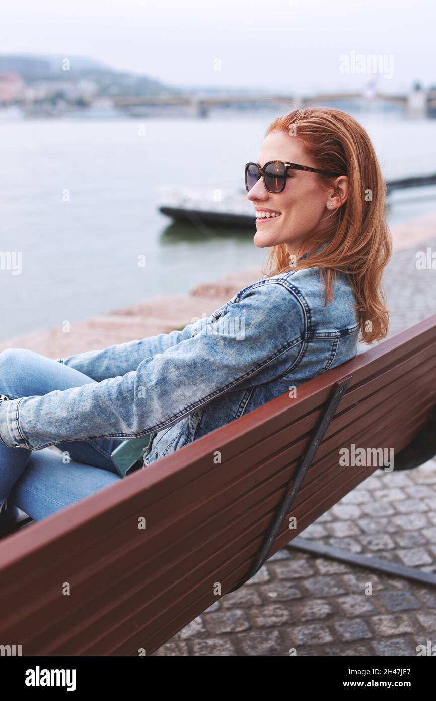 Young balanced redhead Caucasian woman sitting on bench at riverside side view Stock Photo