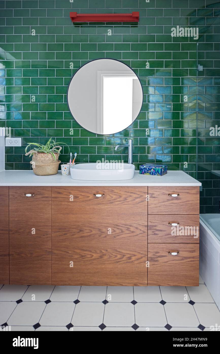 Toilet, wooden cabinet with drawers and white handles, oval sink, green tile wall, white tile floor with small black rhombuses. Red light fixture Stock Photo