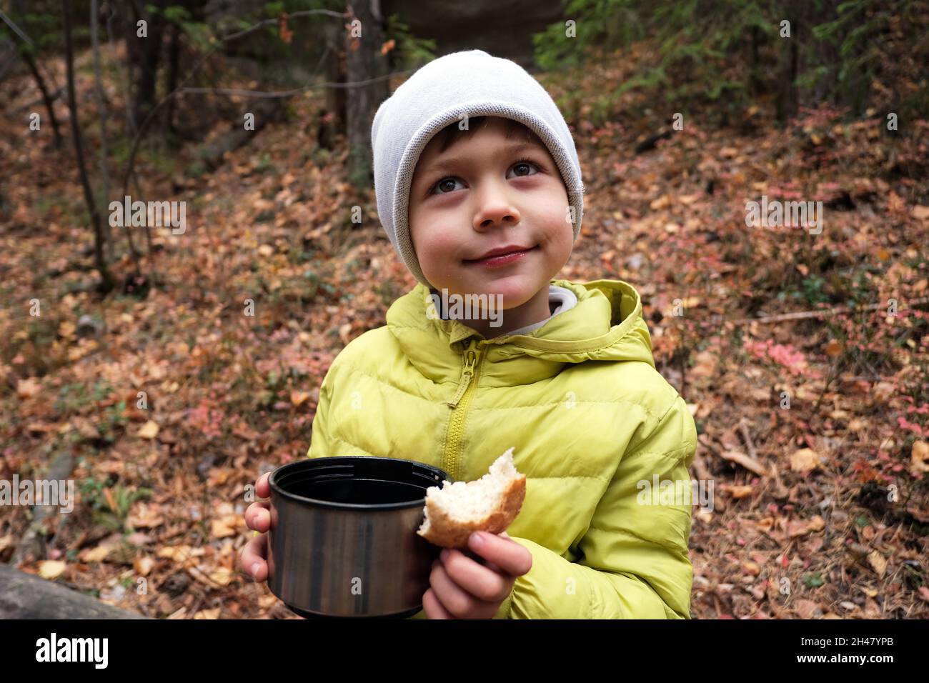 Positive boy in outerwear and cup of hot beverage and bread in hand looking away while sitting in woods during camping Stock Photo