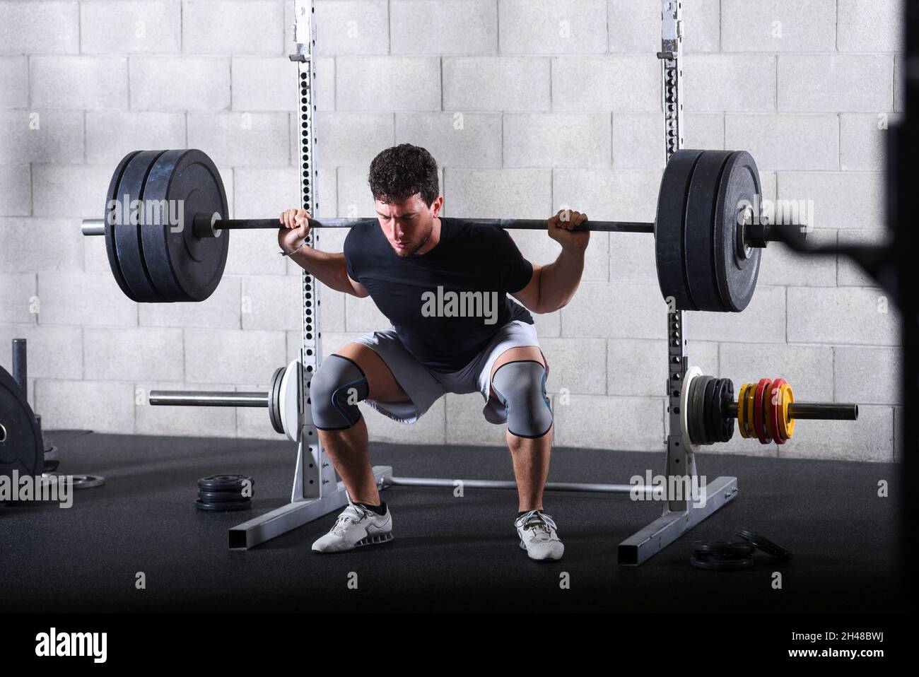Full body view of young man performing back squats exercise in gym with heavy plates. Stock Photo