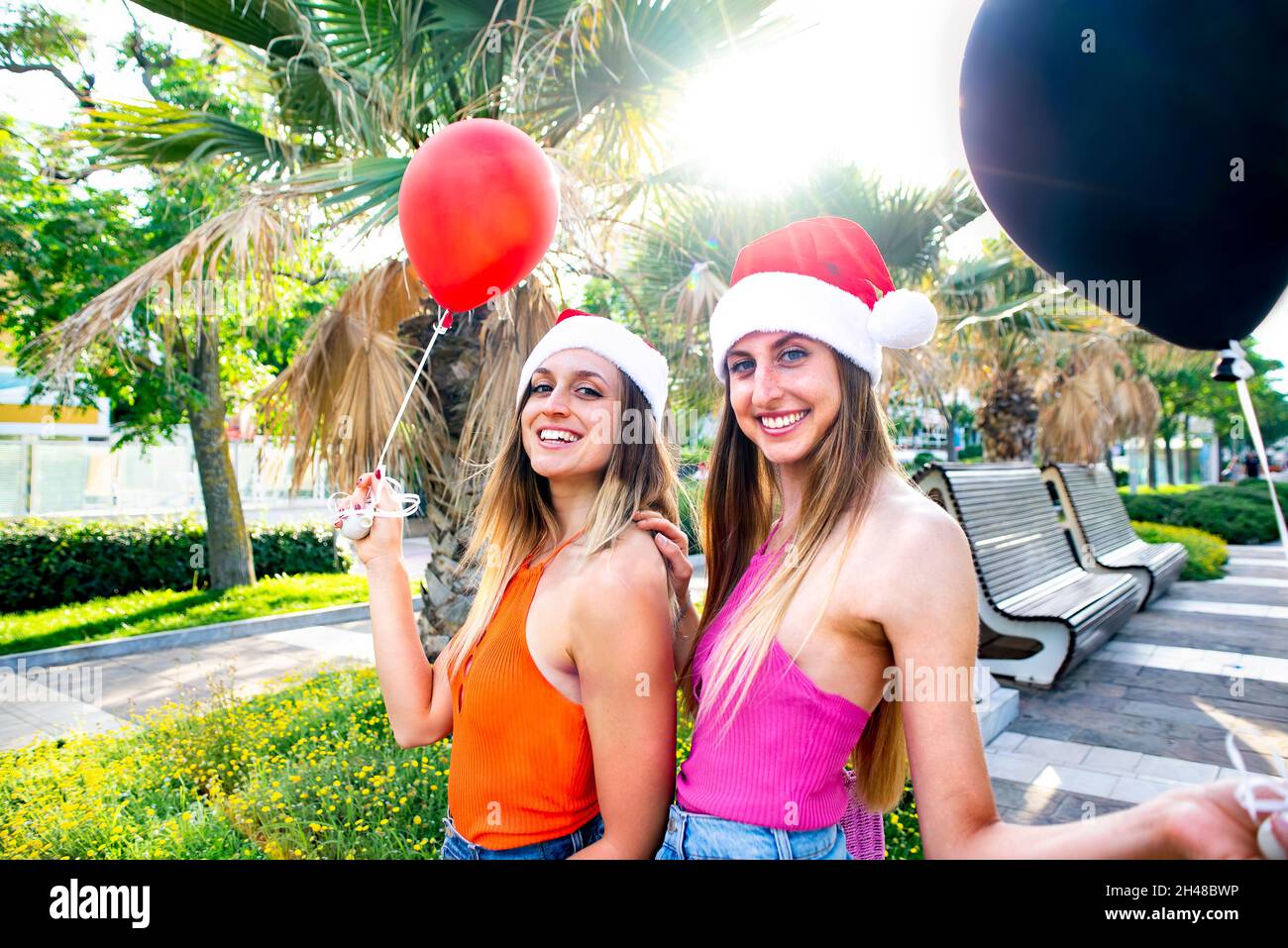 merry christmas! Young adult sisters celebrating xmas wearing a santa claus hat and holding balloons outdoor on winter warm holidays. happy females Stock Photo