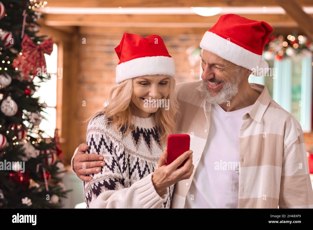 Happy 50 years old couple in santa hats using cell phone on Christmas at home. Stock Photo