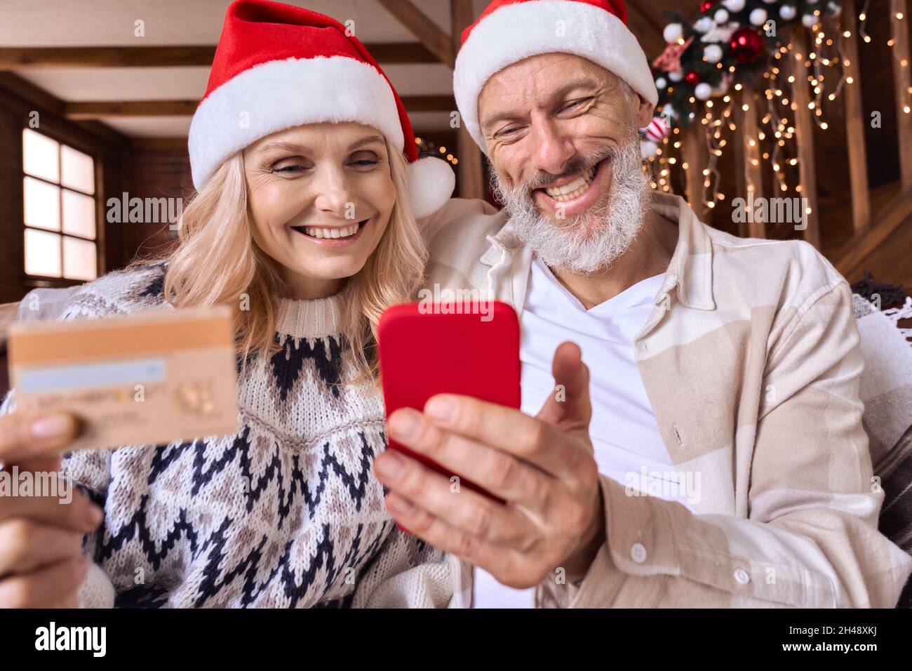 Happy old couple wear santa hats hold phone credit card do Christmas shopping. Stock Photo