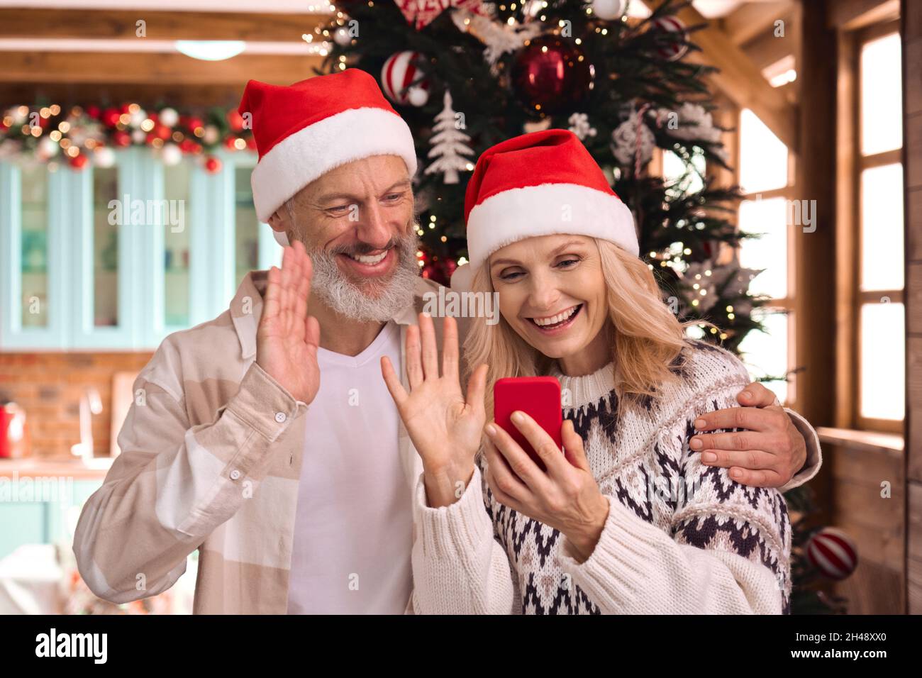 Happy mature couple in santa hats greeting family on phone Christmas video call. Stock Photo