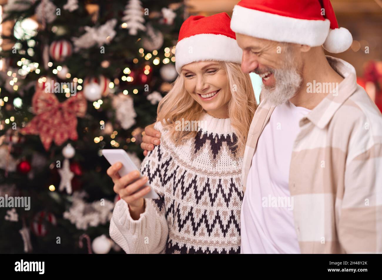 Happy mature couple in santa hats using cell phone near Christmas tree at home. Stock Photo