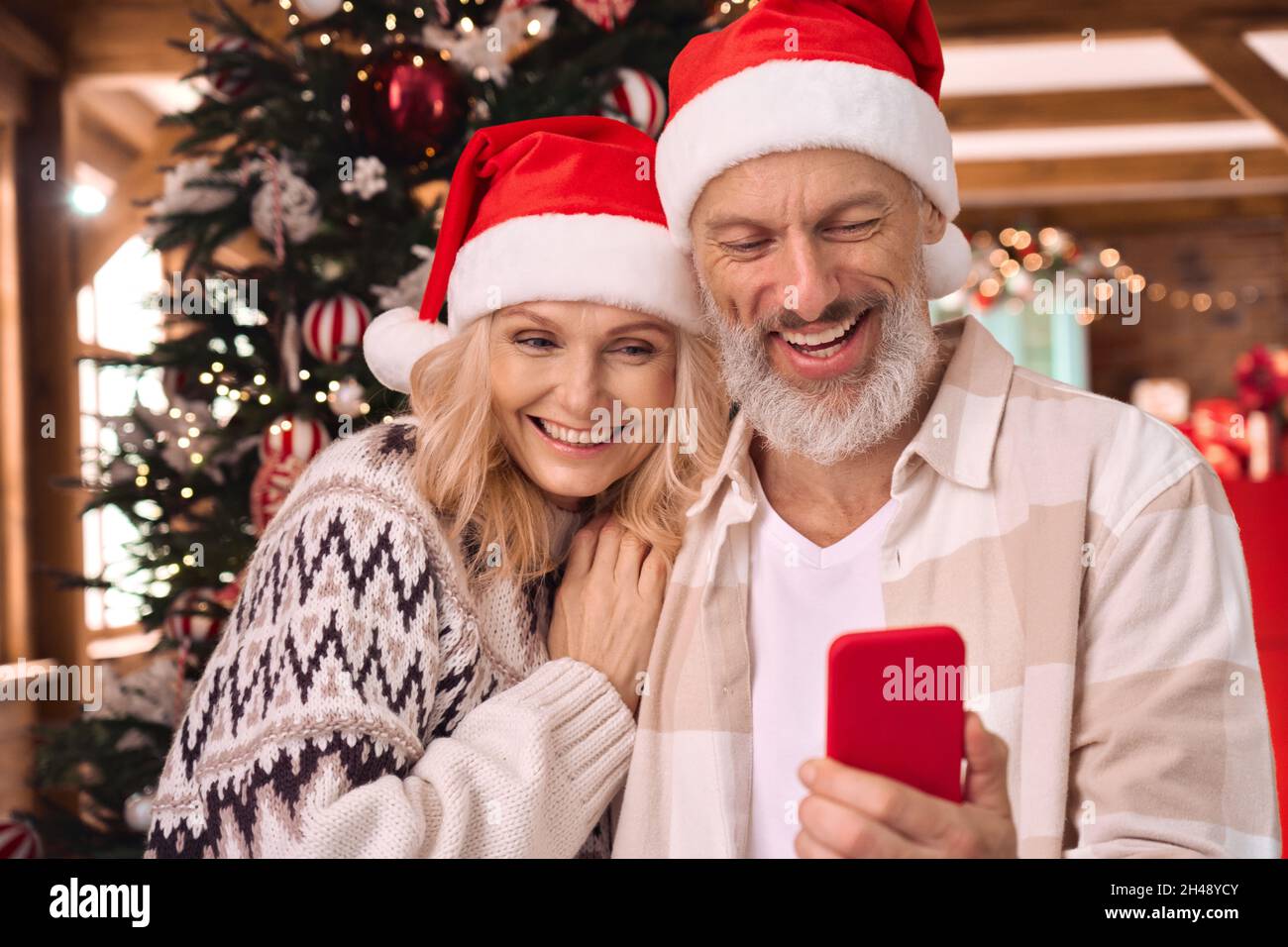 Happy mature couple in santa hats using cell phone near Christmas tree at home. Stock Photo