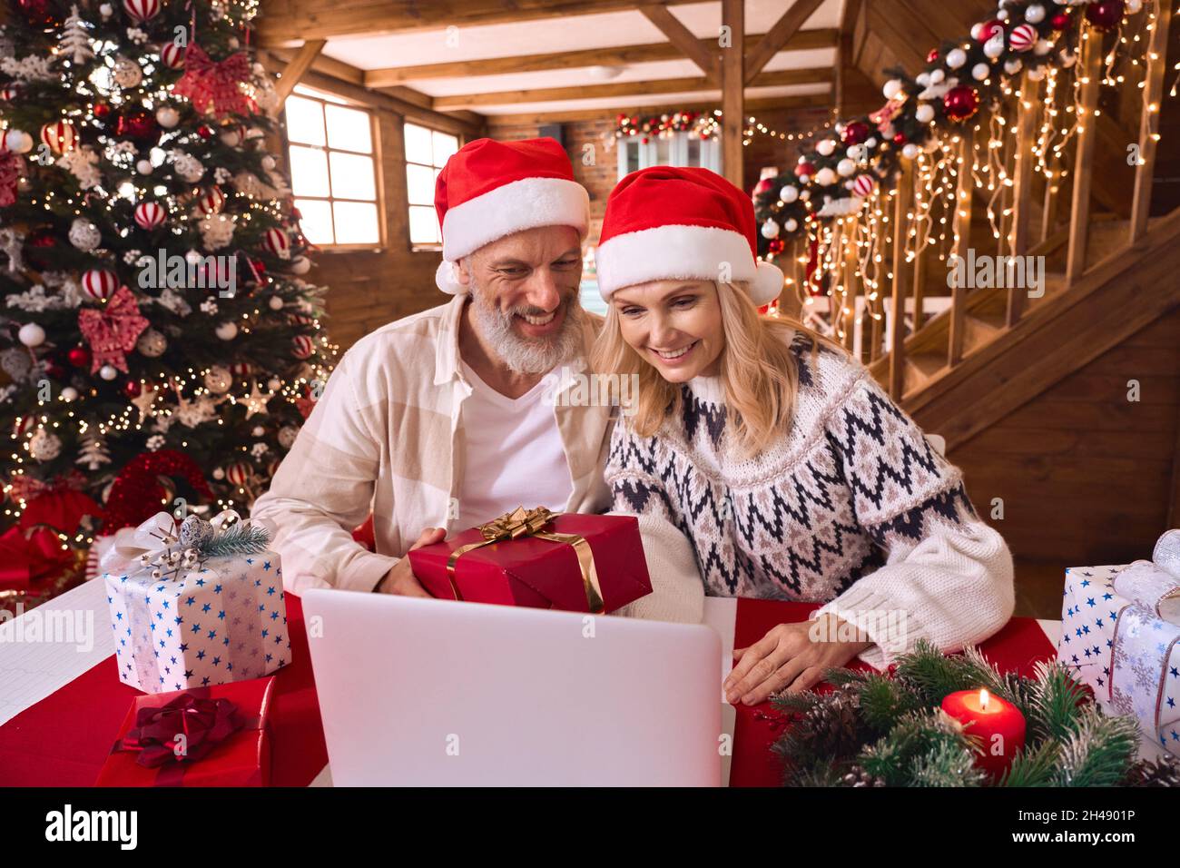 Happy old couple wear santa hats holding gift greeting family on virtual party. Stock Photo