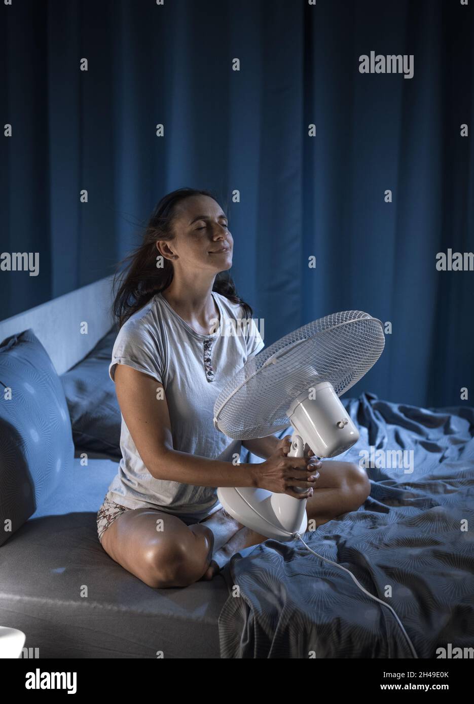 Woman in her bedroom on a hot summer night, she is enjoying fresh air in front of a fan Stock Photo