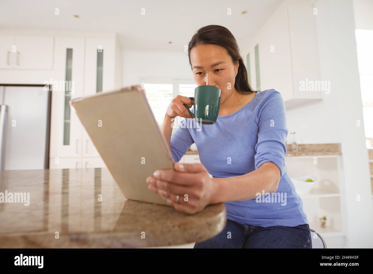 Focused asian woman drinking coffee and using tablet in kitchen Stock Photo