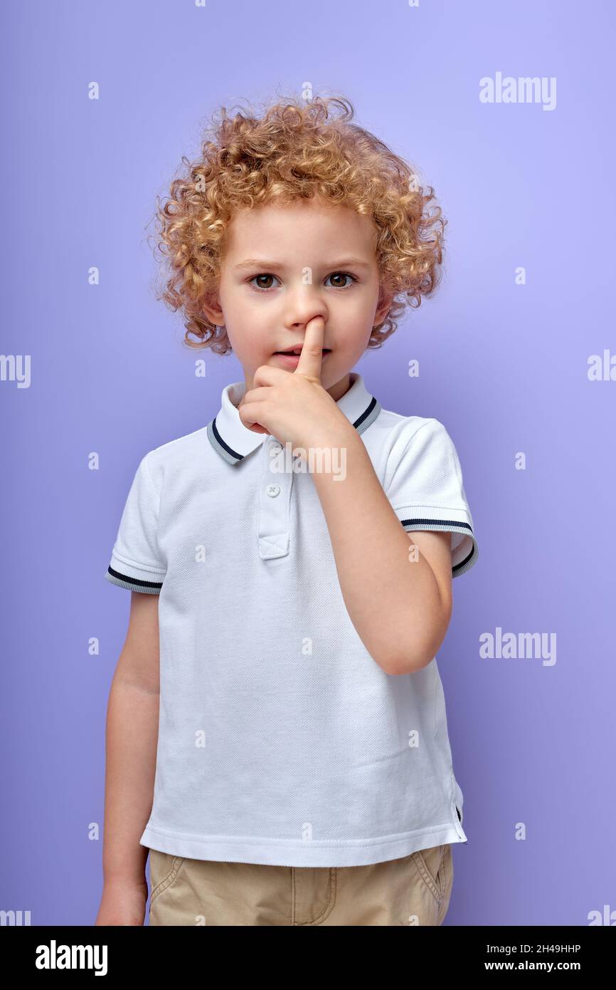 Funny little curly boy picking his nose to explore it and act mischievous for fun childhood and bad habit, isolated over purple studio background, por Stock Photo