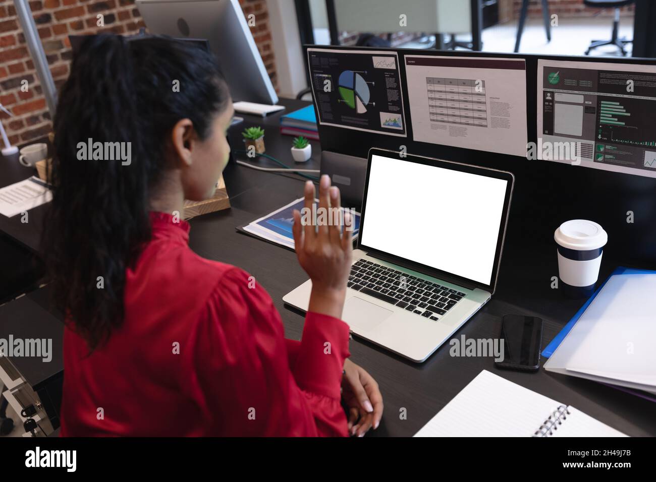 Mixed race woman working in a casual office, sitting at desk, using a laptop computer, waving. Socia Stock Photo