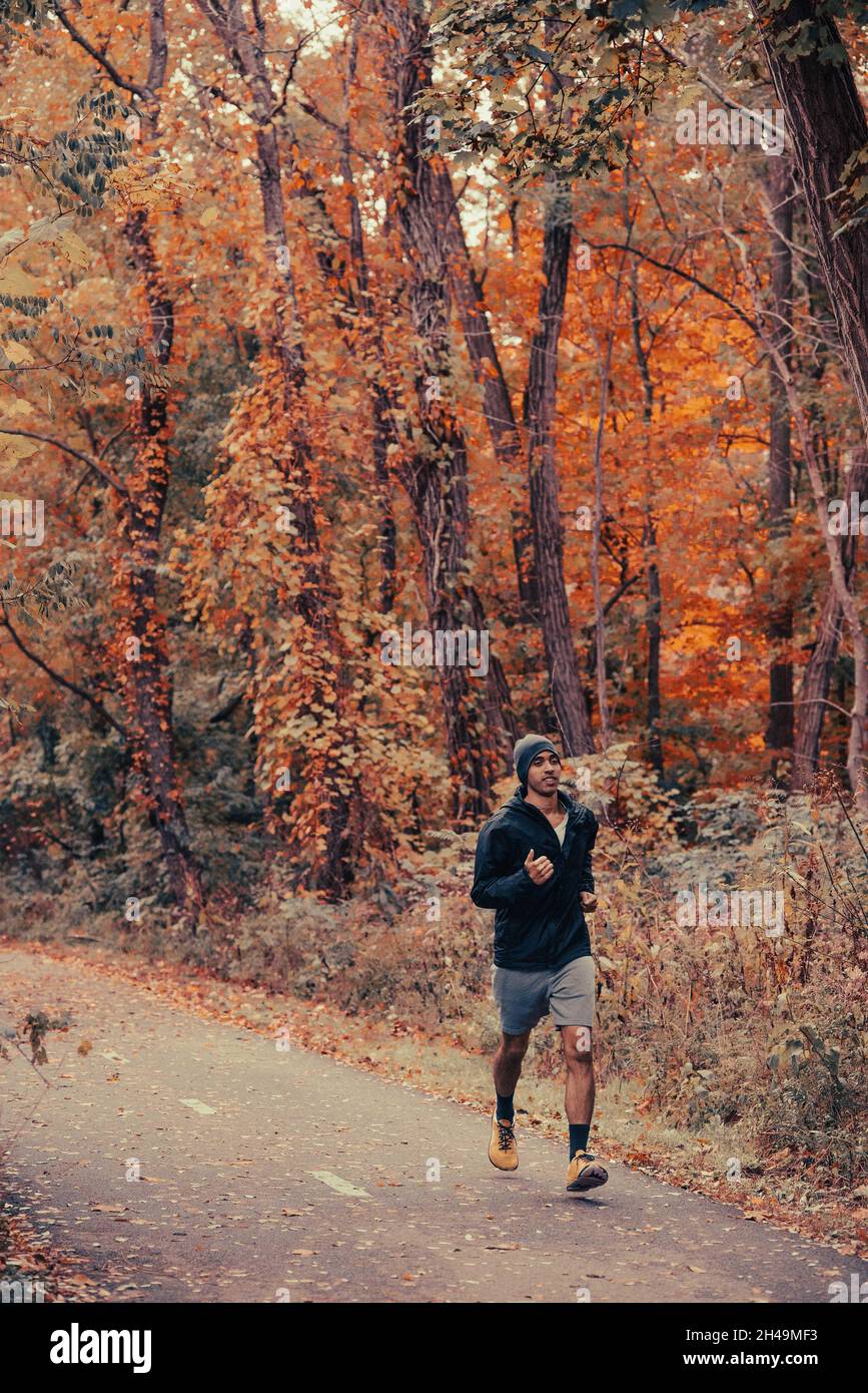 Healthy and fit brown-skinned man runs in the woods on a paved trail in a black jacket and beanie in the fall and autumn season, jogging and training Stock Photo