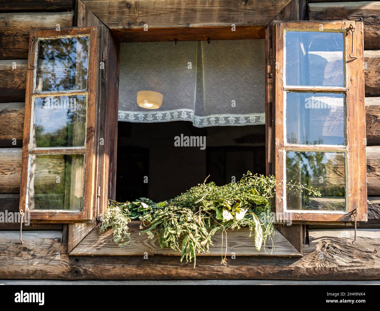 An open window with white curtain, shutters, and drying bunches of herbs, in a traditional, wooden house. Stock Photo