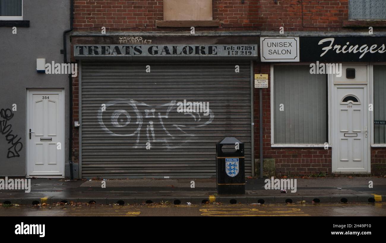 Urban decay, an old shop front shuttered up.  Treats Galore, in Sheffield. Rainy day. Rubbish bin, graffiti. Bleak streetscape. Stock Photo