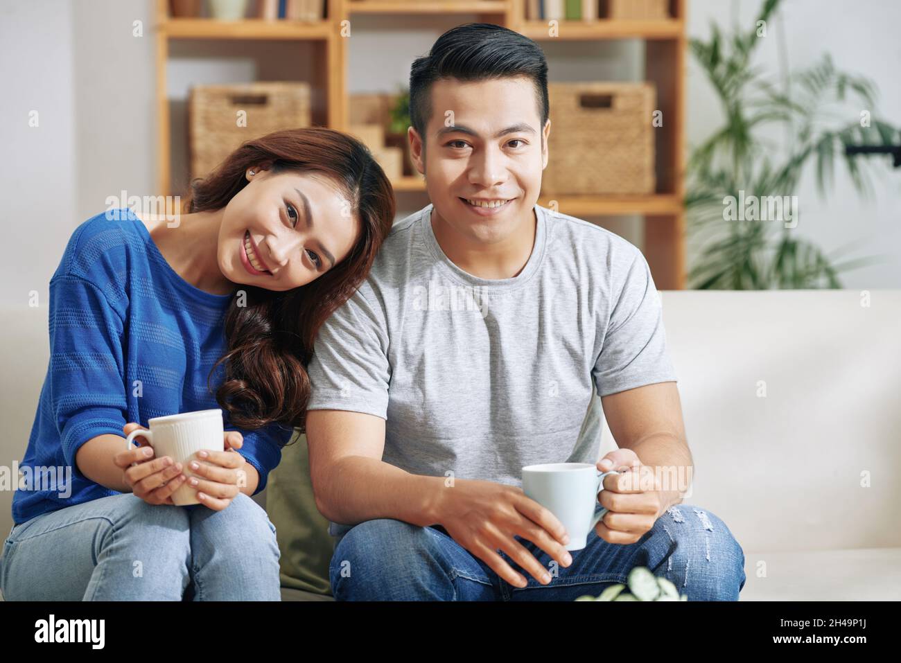 Portrait of smiling young Asian couple in casual outfits sitting on sofa  and drinking coffee from mugs in living room Stock Photo - Alamy