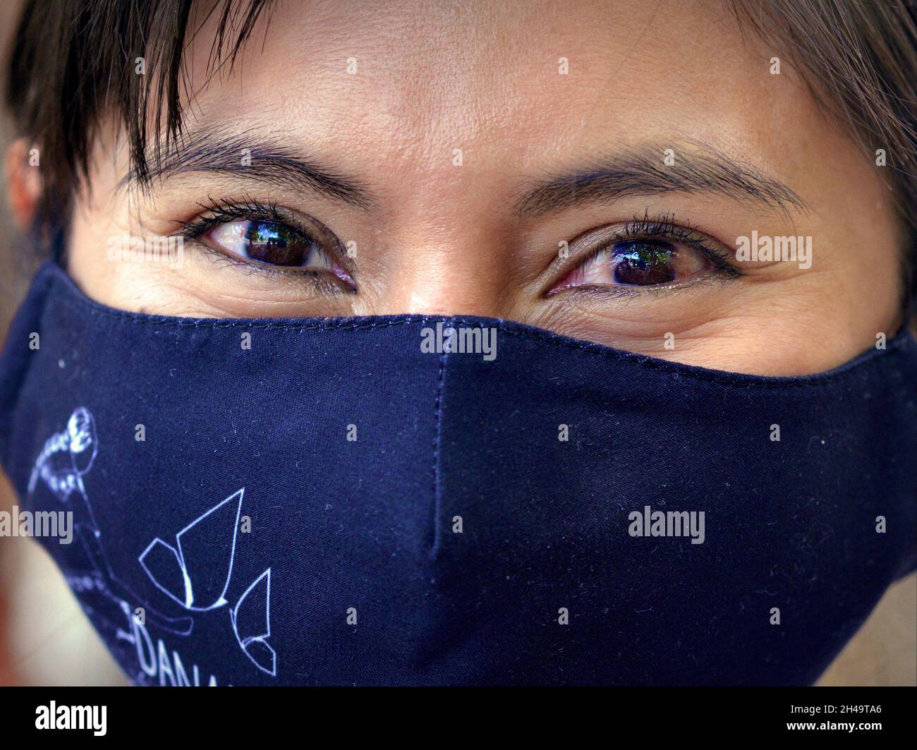 Young Mexican woman with smiling brown eyes wears a black cloth face mask during the global coronavirus pandemic and looks at the viewer. Stock Photo