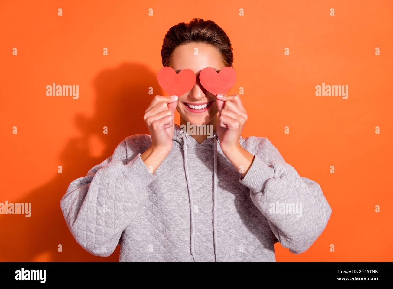 Photo of funny sweet young woman dressed grey hoodie small red hearts cover eyes smiling isolated orange color background Stock Photo