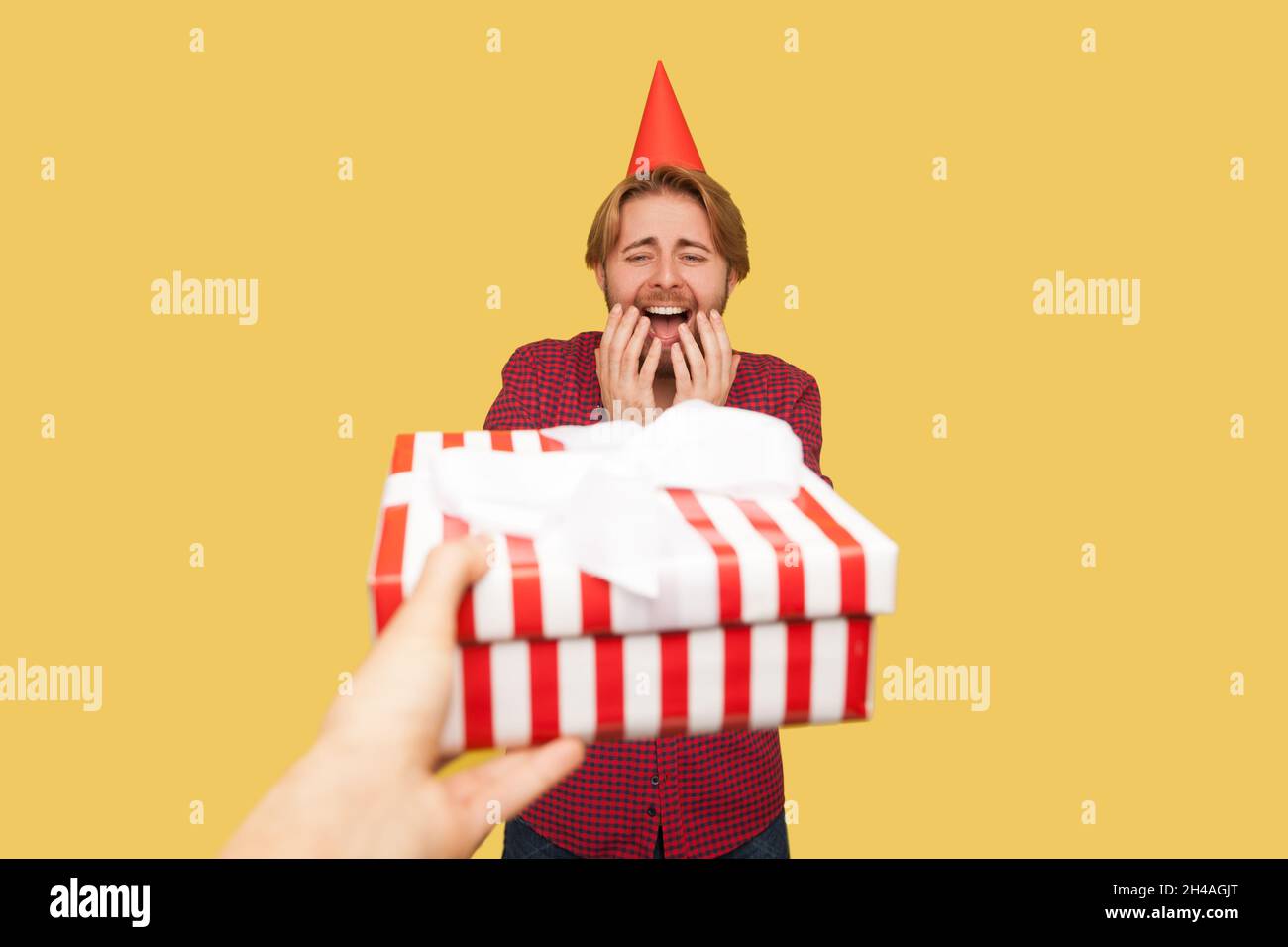 POV hand giving wrapped gift box to guy with red party cone on head, heartwarming man raised arms, can't believe for excellent present. Indoor studio shot isolated on yellow background. Stock Photo