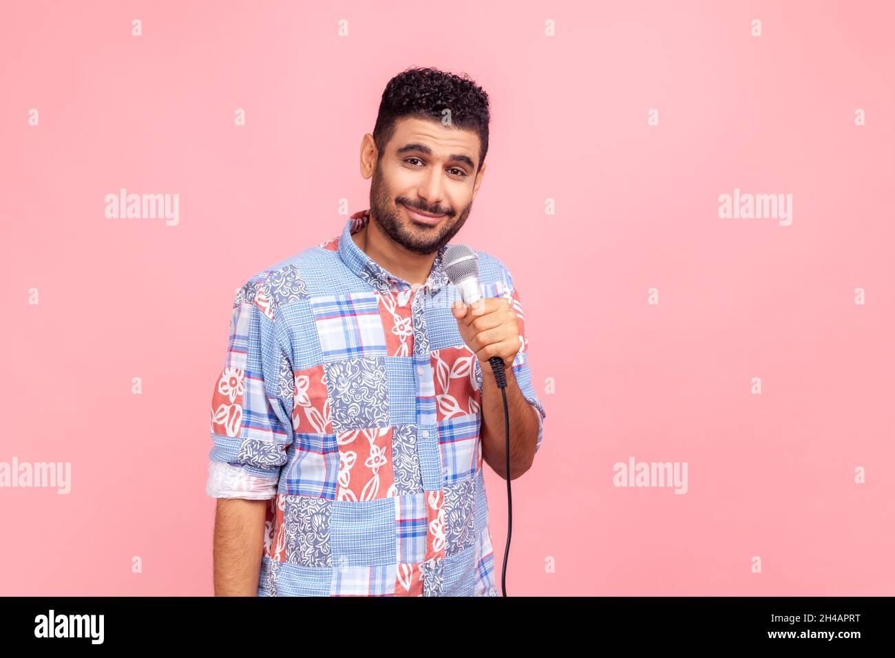 Handsome satisfied man with beard holding microphone in hands, taking interview, looking at camera with pleasant smile, wearing blue shirt. Indoor studio shot isolated on pink background. Stock Photo