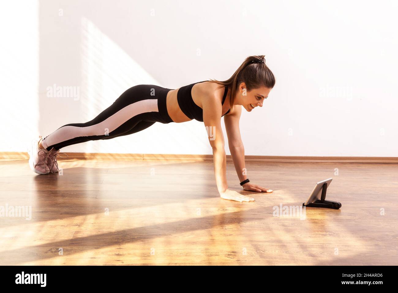 Woman doing plank exercises while watching video on tablet, learning online, repeat after coach, wearing black sports top and tights. Full length studio shot illuminated by sunlight from window. Stock Photo