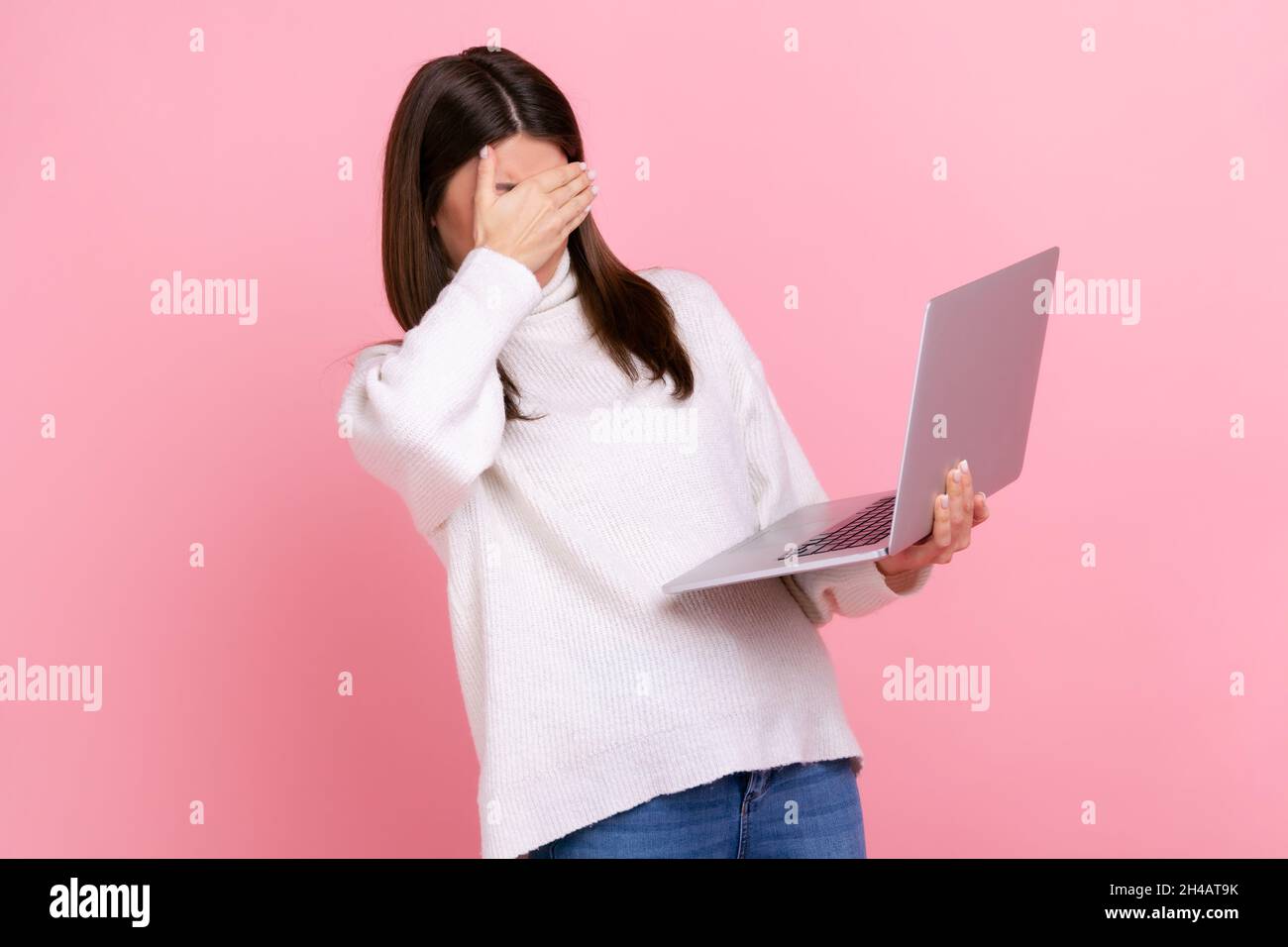 Portrait of brunette female holding laptop computer, covering eyes with palm, sees disgust content, wearing white casual style sweater. Indoor studio shot isolated on pink background. Stock Photo