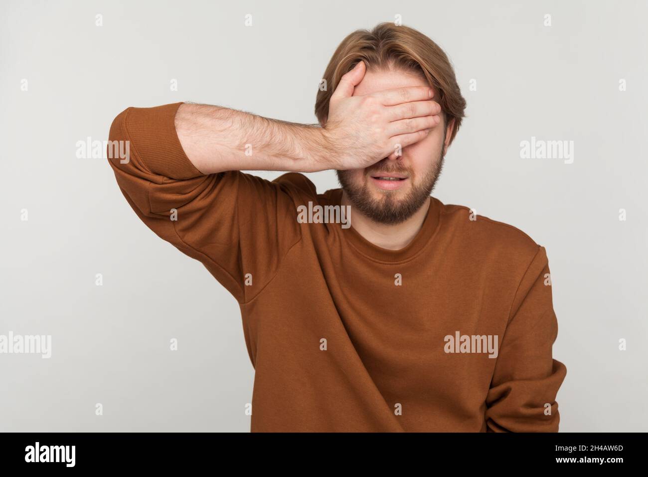 Portrait of man with beard wearing sweatshirt, closing eyes with hand, dont want to see that, ignoring problems, hiding from stressful situations. Indoor studio shot isolated on gray background. Stock Photo