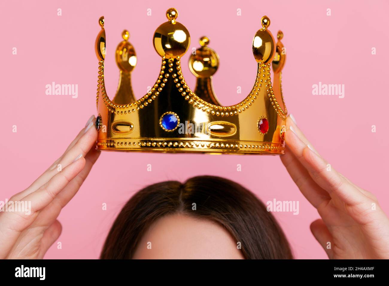 Unknown woman putting on golden crown, arrogance and privileged status, concept of self confidence in success, self-motivation and dreams to be best. Indoor studio shot isolated on pink background. Stock Photo
