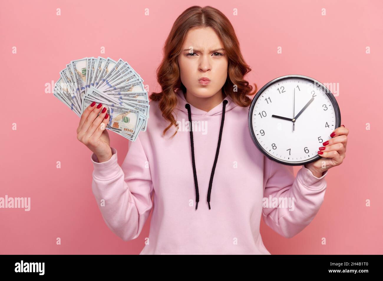 Portrait of angry displeased curly haired teen girl in hoodie holding dollar banknotes and wall clock, looking strictly at camera. Indoor studio shot isolated on pink background Stock Photo