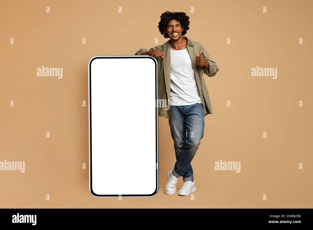 Hnadsome Smiling Black Guy Leaning On Big Smartphone With Blank White Screen And Showing Thumb Up, Positive African American Male Recommending New Mob Stock Photo